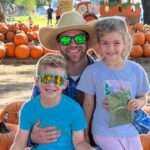 Family at a pumpkin patch posing for a photo; man with straw hat and sunglasses flanked by two kids among stacked pumpkins, festive cartoon pumpkin decorations around them