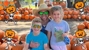 Family at a pumpkin patch posing for a photo; man with straw hat and sunglasses flanked by two kids among stacked pumpkins, festive cartoon pumpkin decorations around them