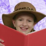 Girl wearing a wide-brimmed hat, smiling while reading a red book against a purple background.