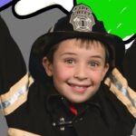 Smiling child in a firefighter costume with a helmet labeled 'Fire Chief', arms raised in celebration on a colorful backdrop.
