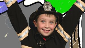 Smiling child in a firefighter costume with a helmet labeled 'Fire Chief', arms raised in celebration on a colorful backdrop.