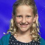 Smiling blonde girl with wavy hair in a polka-dot navy shirt and teal cardigan, posed against a blue starry backdrop.