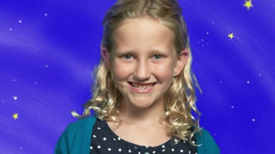 Smiling blonde girl with wavy hair in a polka-dot navy shirt and teal cardigan, posed against a blue starry backdrop.
