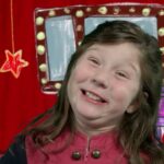Young girl smiling on a stage with a red curtain and drawn props behind her.