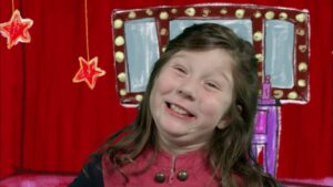 Young girl smiling on a stage with a red curtain and drawn props behind her.