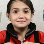 A smiling young girl in a red jacket sits at a space station mockup with control panels on the wall to the left and a starry window on the right.