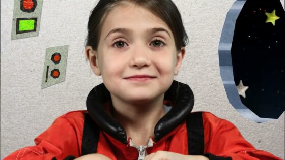 A smiling young girl in a red jacket sits at a space station mockup with control panels on the wall to the left and a starry window on the right.