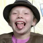Girl laughing with tongue out, wearing a hat and brown vest, in front of a colorful drawn backdrop.