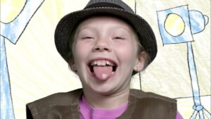 Girl laughing with tongue out, wearing a hat and brown vest, in front of a colorful drawn backdrop.