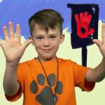 Smiling boy in an orange shirt with a gray paw print raises both hands; a red stop-hand sign sits on a post behind him against a blue-painted backdrop.