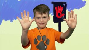 Smiling boy in an orange shirt with a gray paw print raises both hands; a red stop-hand sign sits on a post behind him against a blue-painted backdrop.
