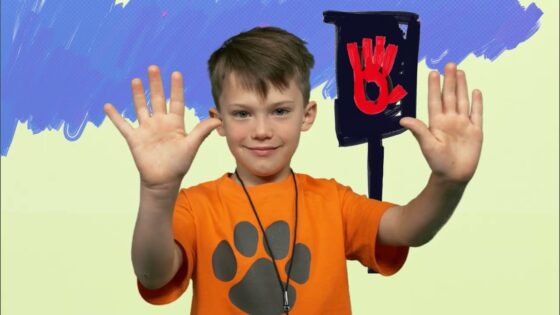 Smiling boy in an orange shirt with a gray paw print raises both hands; a red stop-hand sign sits on a post behind him against a blue-painted backdrop.