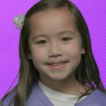 Smiling young girl with long hair and a white flower in her hair, wearing a purple top, against a purple background