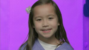 Smiling young girl with long hair and a white flower in her hair, wearing a purple top, against a purple background