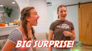 Thumbnail showing two women indoors reacting excitedly to a surprise, one with braided hair in a gingham top and the other in a gray t‑shirt, with 'BIG SURPRISE!' text across the bottom.