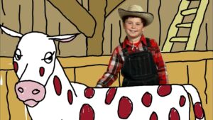 Smiling boy in a plaid shirt, overalls and a straw hat stands beside a white cow with red spots in a barn.