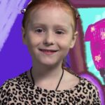 Smiling young girl with red hair wearing a leopard-print shirt, posed in front of a bright, cartoon-style purple background.