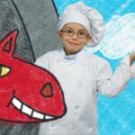 Child in a white chef uniform and hat stands beside a large red cartoon dragon drawing on a blue background, smiling at the camera.