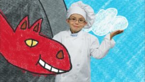 Child in a white chef uniform and hat stands beside a large red cartoon dragon drawing on a blue background, smiling at the camera.