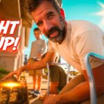 Bearded man crouching outdoors, pointing a lighter to a small flame, with bold 'LIGHT IT UP!' text on the left and a sunny sky in the background.