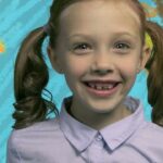 Girl with brown pigtails and a light purple shirt smiles at the camera against a bright painted backdrop.