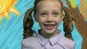 Girl with brown pigtails and a light purple shirt smiles at the camera against a bright painted backdrop.