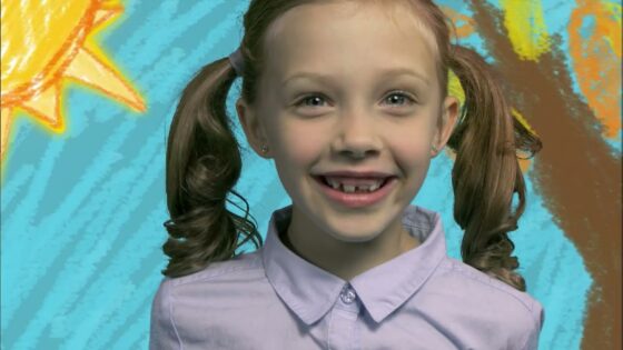 Girl with brown pigtails and a light purple shirt smiles at the camera against a bright painted backdrop.