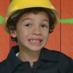 Young child in a yellow construction helmet giving a thumbs up, standing in front of a wooden backdrop.