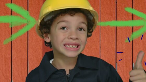 Young child in a yellow construction helmet giving a thumbs up, standing in front of a wooden backdrop.