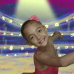 Young girl in a pink leotard posing and smiling while dancing on a bright stage backdrop with pink curtains and spotlights.