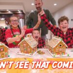 Joyful family posing with decorated gingerbread houses on a kitchen table during Christmas, a man stands behind them applauding as kids cheer.