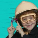 Smiling child wearing a hard hat and safety goggles, pointing at a cartoon crab on their finger against a turquoise background.