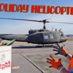 Volunteer in a blue hard hat and orange vest beside a U.S. Army helicopter on a tarmac, with a Toys for Tots box in front.