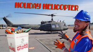 Volunteer in a blue hard hat and orange vest beside a U.S. Army helicopter on a tarmac, with a Toys for Tots box in front.