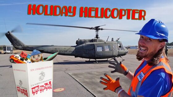 Volunteer in a blue hard hat and orange vest beside a U.S. Army helicopter on a tarmac, with a Toys for Tots box in front.
