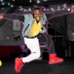 DJ Dave leaps in a dim party room with colorful string lights; wearing a yellow hoodie, denim vest, white and black pants, red sneakers; DJ Dave logo in corner.