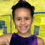 Smiling young girl in a purple dress raises her right index finger against a bright yellow background with blue accents.