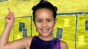 Smiling young girl in a purple dress raises her right index finger against a bright yellow background with blue accents.