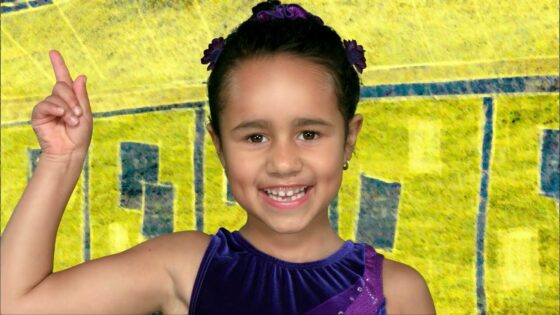 Smiling young girl in a purple dress raises her right index finger against a bright yellow background with blue accents.