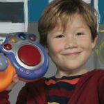 Smiling boy holds a colorful toy with an orange ring and purple buttons toward the camera.
