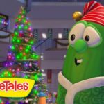 Cheerful green VeggieTales character wearing a red striped hat and scarf beside a lit Christmas tree in a snowy mall scene