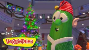 Cheerful green VeggieTales character wearing a red striped hat and scarf beside a lit Christmas tree in a snowy mall scene