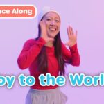 Smiling woman in a red sweatshirt waves with a purple background and the bold blue 'Joy to the World' text overlay; top-left 'Dance Along' badge present.