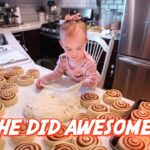 Two-year-old girl with a pink bow stretches dough among many cinnamon rolls on a kitchen island, a baking scene with a fridge in the background, overlay text says 'SHE DID AWESOME!'