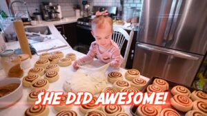 Two-year-old girl with a pink bow stretches dough among many cinnamon rolls on a kitchen island, a baking scene with a fridge in the background, overlay text says 'SHE DID AWESOME!'