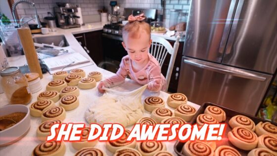 Two-year-old girl with a pink bow stretches dough among many cinnamon rolls on a kitchen island, a baking scene with a fridge in the background, overlay text says 'SHE DID AWESOME!'