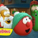 Three VeggieTales characters—a red tomato wearing a green knit cap, a green cucumber in a red plaid hat and vest, and a Santa-like veggie—smile in a snowy town square with warm lights in the background.