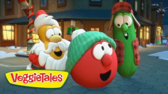 Three VeggieTales characters—a red tomato wearing a green knit cap, a green cucumber in a red plaid hat and vest, and a Santa-like veggie—smile in a snowy town square with warm lights in the background.