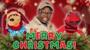 Person in safari-style outfit with two puppets, 'Merry Christmas' text in front, festive red background with pine garland and pinecones.