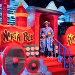 Colorful ice sculpture train labeled 'North Pole Express' with a man and two children posing on red steps in winter coats.